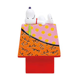 A decorative figurine of Snoopy lying on top of a colorful dog house. Snoopy is white with a small purple nose, wearing a red collar.