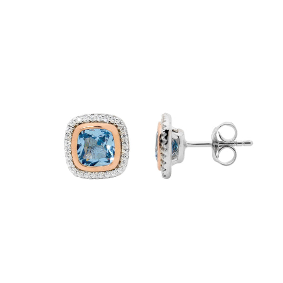 A pair of two-tone stud earrings with a cushion cut blue center stone, surrounded by a rose gold plated bezel and clear cubic zirconia halo, set in sterling silver.